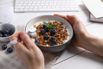 Woman holding bowl of tasty granola with blueberries at white wooden table, closeup