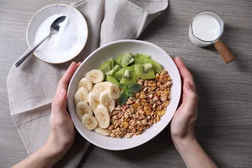Plexiglas foto achterwand Woman holding bowl of tasty granola with banana and kiwi at grey wooden table, top view © New Africa