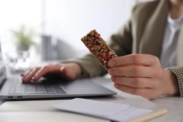 Poster Woman holding tasty granola bar working with laptop at light table in office, closeup © New Africa