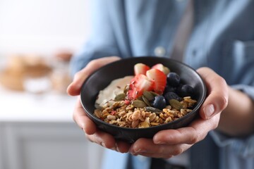 Woman holding bowl of tasty granola with berries, yogurt and seeds indoors, closeup