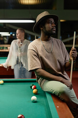 Side view portrait of Black adult man wearing hat posing on billiards table in bar