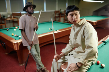 Portrait of Asian man sitting on pool table and looking away from camera in bar, copy space