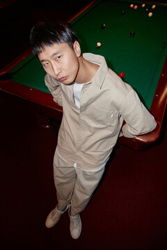 Vertical full length portrait of Asian man standing by pool table and looking up at camera shot with flash