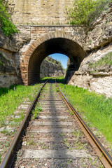 View of the rails going into the distance through an old railway tunnel made of stone between the rocks.