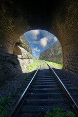 From the railway tunnel, a view of the end of the tunnel and the rails going into the distance between two rocks against the sky