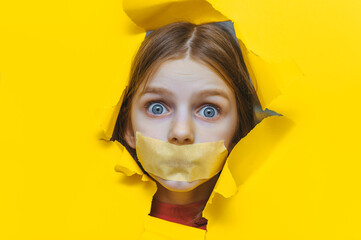 A child, his mouth covered with masking tape, peers through a torn hole in yellow paper. The concept of a ban on opinion, unwillingness to listen to children, restriction of freedom of speech.