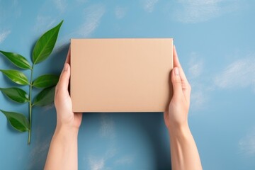 Person holding a plain brown cardboard box against a vibrant blue background with green leaves. Hands Holding Brown Cardboard Box on Blue
