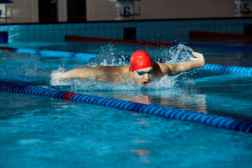 Young man in red cap and goggles, swimmer in motion, showing butterfly stroke, training, swimming...