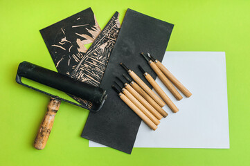 Linocut tools. Cutters, roller, black linoleum and white sheet of paper on a green background. Handicraft and design concept. Handmade creativity.
