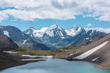 Closeup of most beautiful turquoise alpine lake against snow-covered range with few pointy peaks. Azure water of mountain lake close-up against three snowy peaked tops under clouds in cloudy blue sky.