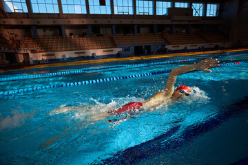 Young man, swimmer in red cap and goggles in motion, training freestyle stroke, swimming in pool...