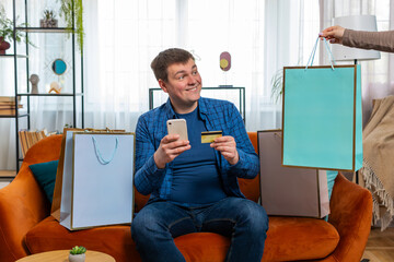 Happy shopaholic consumer man sitting with shopping bags at apartment making online order, payment...