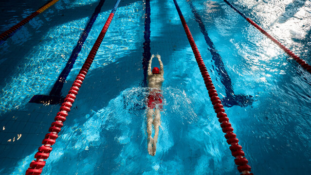 Top view of muscular, athletic young man, swimmer in red cap in motion, showing strength, training, swimming in pool indoors. Concept of professional sport, health, endurance, active lifestyle
