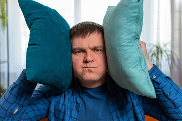 Repair work at neighbors. Irritated angry fat man covers ears with cushions pillows annoyed by...