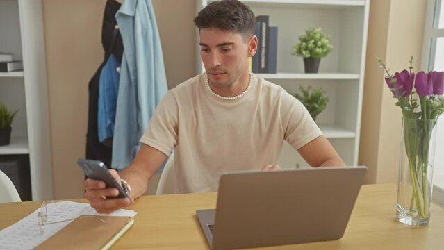 A handsome young hispanic man checks his smartphone while working on a laptop in a modern home interior.