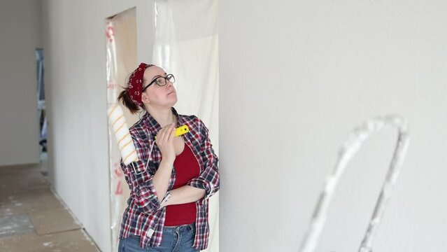 A woman with glasses and a roller in her hands carefully examines a white wall getting ready to paint. High quality FullHD footage