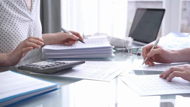 Business colleagues, accountants, or auditors are reviewing financial data in fair office. Two professionals are analyzing paper reports and using calculators, focusing on aspects of audit and taxes