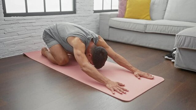 A fit hispanic man practices yoga on a mat in a modern living room, exhibiting wellness and tranquility.