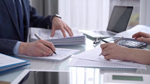 Business colleagues, accountants, or auditors are reviewing financial data in fair office. Two professionals are analyzing paper reports and using calculators, focusing on aspects of audit and taxes