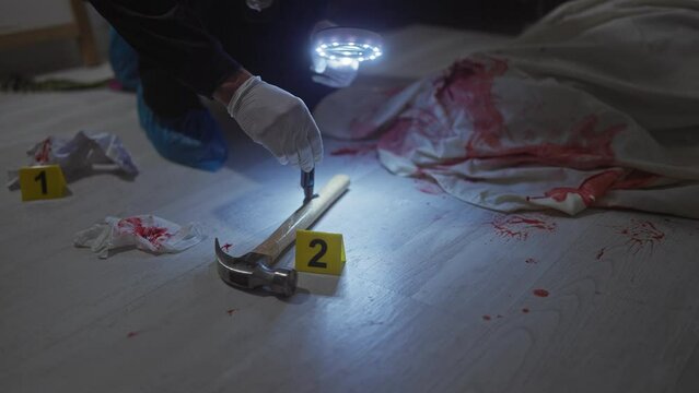Gloved hand examining hammer at a bloody crime scene indoors with evidence markers and forensic light.