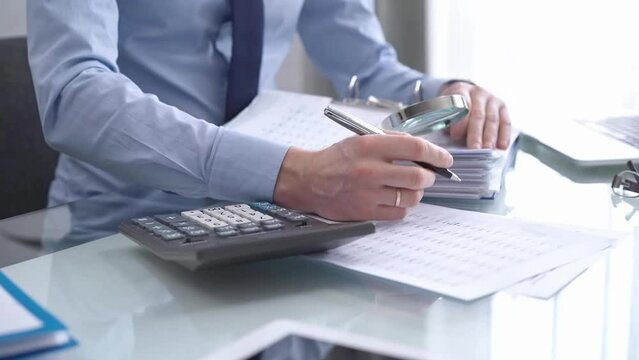 A businessman is using a calculator and a magnifying glass to analyze financial documents at his glass desk in a modern and fair office setting, focusing on aspects of audit and taxes in business