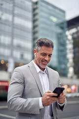 Smiling middle aged business man wearing earbud holding phone having call walking on city street. Mature businessman with earphone using smartphone listening podcast in app on cellphone outdoors. - 783155631