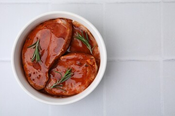 Raw marinated meat and rosemary in bowl on white tiled table, top view. Space for text