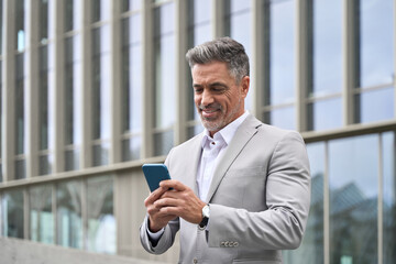 Happy elegant middle aged business man ceo, 45 years old businessman leader investor executive wearing suit holding cellphone using mobile cell phone standing outdoors on city street background. - 783160677