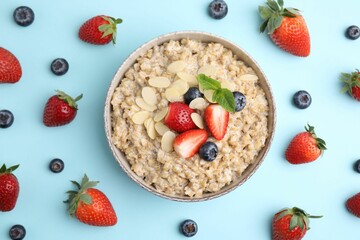Tasty oatmeal with strawberries, blueberries and almond flakes in bowl surrounded by fresh berries on light blue background, flat lay