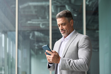 Successful rich middle aged businessman using cellphone, busy older business man leader investor, mature male executive in suit holding mobile phone standing at office window looking at smartphone. - 783162015