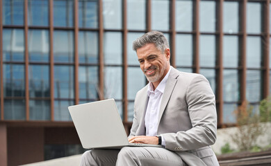 Happy busy middle aged business man working on laptop outside office. Smiling older professional businessman executive ceo manager wearing suit sitting outdoors using computer managing data. - 783162265