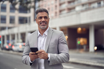 Smiling middle aged business man using smartphone looking away outdoors. Happy mature businessman ceo entrepreneur investor wearing suit standing outside office holding mobile cell phone in hands. - 783164495