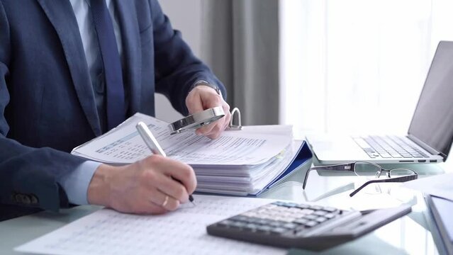 Businessman reviewing ring folder of financial documents with magnifying glass at desk in modern office. Audit and taxes in business