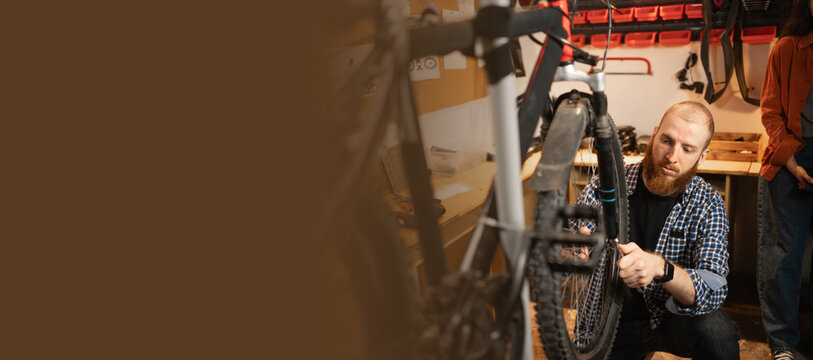 Young handsome beard bicycle mechanic repairing bicycles in a workshop or garage. Banner. - Powered by Adobe