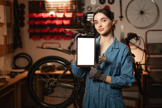 Young woman repairing bicycle in workshop, holding digital tablet with Mockup image