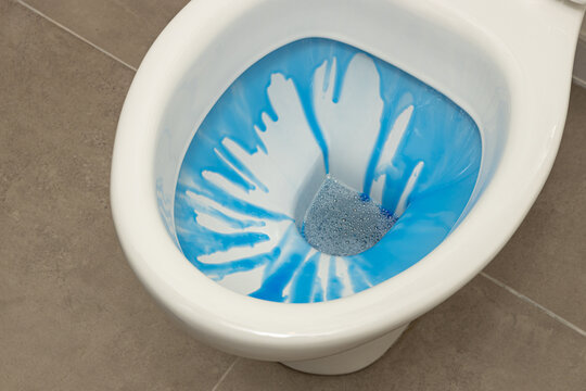 Close up concept shot of a toilet bowl with blue toilet cleaner running down into the water with bubbles; close up of modern toilet bowl being cleaned with toilet detergent, with grey tile floor.