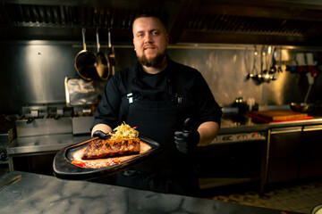 in a professional kitchen the chef stands in his hands with a prepared dish and smiles at the camera