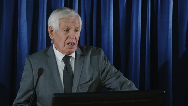 Senior politician in formal suit standing at lectern against blue curtain and speaking in microphone during debates or press conference