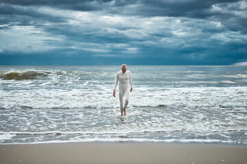 Hairless girl with alopecia in white futuristic suit comes out of cold restless sea on sandy beach,...