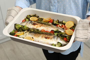 Woman holding baking dish with delicious fish and vegetables indoors, closeup