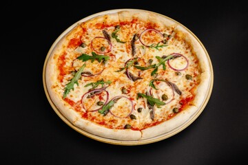 Pizza with anchovies, red onion, capers, and arugula on a black background.