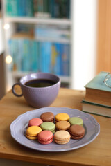 Purple plate filled with pastel macarons, cup of tea or coffee, vintage books and reading glasses on the table. Colorful bookcase in the background. Selective focus.