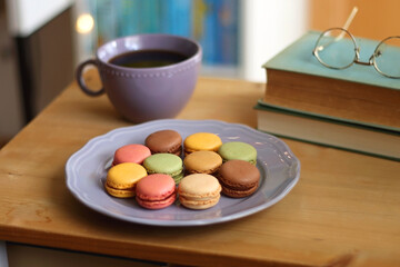 Purple plate filled with pastel macarons, cup of tea or coffee, vintage books and reading glasses on the table. Colorful bookcase in the background. Selective focus.