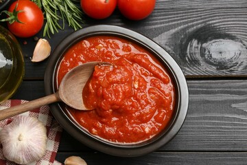 Homemade tomato sauce in bowl, spoon and fresh ingredients on black wooden table, flat lay