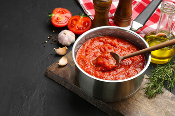 Homemade tomato sauce in pot, spoon and ingredients on dark table, space for text