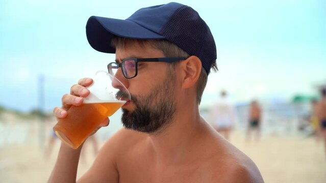 Man drinks beer from glass on background of sea beach and people playing volleyball. Vacation, travel, holiday.