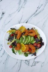 Gourmet leaves salad. Mediterranean flavor. Top view of colorful salad with lettuce, cabbage, tomatoes, fried shrimps, avocado and orange, served on a white dish on the white marble table.	
