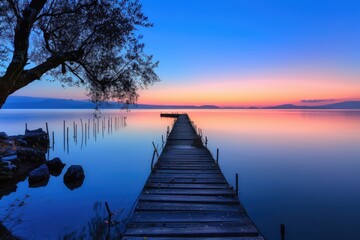 Wooden pier extending into a tranquil sunset - The old wooden pier leads the eye into the infinite horizon, under the splendid colors of a tranquil sunset over calm waters - obrazy, fototapety, plakaty