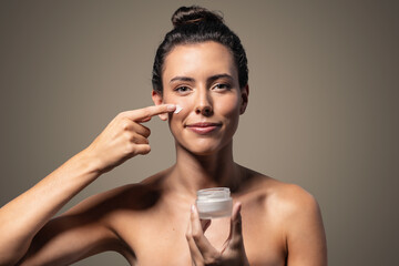 Beautiful young woman caring of her skin while putting on cream looking at camera on isolated brown background.