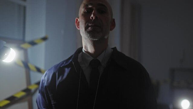 A stern bald man displays a police badge in a dimly lit indoor crime scene, suggesting investigation and authority.